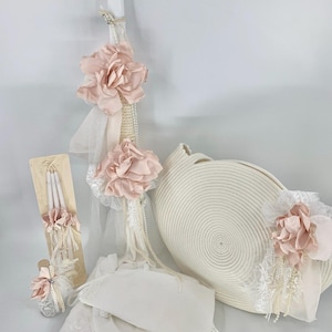 May include: A white christening set featuring a white candle adorned with pink flowers and lace. The set includes a white basket, a white gown, and a white candle holder.