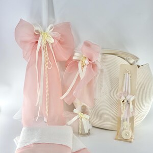 May include: A set of baby christening gifts, including a white candle with a pink bow, a small white basket with a pink bow, a white towel with a pink border, a small bottle of lotion with a pink bow, and a small white candle with a pink bow.