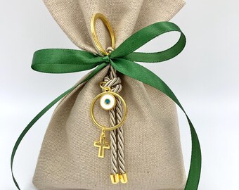 Op de afbeelding: Een klein, beige stoffen tasje met een groen lint dat eromheen is gebonden. Een gouden hanger met een blauw boze oog en een kruis hangt aan het tasje.