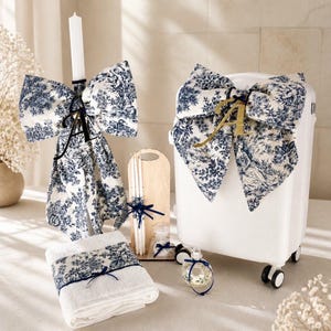May include: A white suitcase and a tall candle, both adorned with large bows made of blue and white toile fabric. The suitcase features a gold letter A, while the candle has a black letter A. A folded white towel with a matching toile band and a small glass bottle are included.