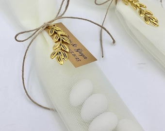 May include: Small white mesh bags tied with twine, each adorned with a gold leaf charm and a small tag. The bags contain white candies. The tag reads "Irini & Gregor 05-2025".