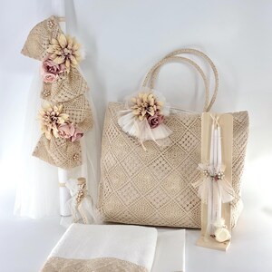 May include: A set of baptism gifts including a white candle with a crocheted lace and pink flower embellishment, a crocheted tote bag with a pink flower embellishment, a white towel with crocheted lace trim, and a white candle with a crocheted lace and pink flower embellishment.