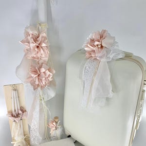 May include: A selection of white and pale pink baptism or wedding decorations. Features a decorated candle, suitcase, candles, and a small bottle, all embellished with lace, ribbons, and floral details. The overall theme is elegant and celebratory.