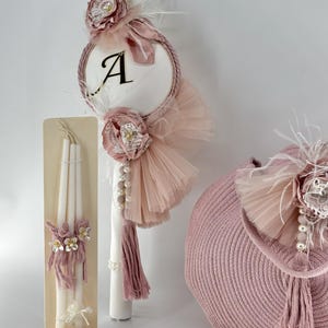 May include: A pink and white christening candle set with a large pink and white tulle bow, a small wooden stand with three white candles, and a pink woven basket with a pink tulle bow and a white feather.