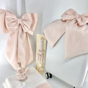 May include: A white suitcase with pink satin bows, a white towel with a pink border, a white candle with a pink satin bow, a small bottle with a pink bow, and a white card with a white candle and pink ribbon.