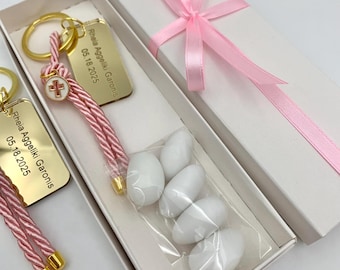 May include: A gift set featuring a gold-colored keychain with a pink and white cord, a cross charm, and a personalized tag. The set includes a box with white sugared almonds and a pink ribbon.