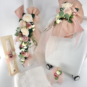May include: A selection of baptism or wedding decorations. Features a white towel with a pink lace trim, candles, and a decorated suitcase. The candles and suitcase are adorned with pink and white roses, greenery, and sheer fabric.