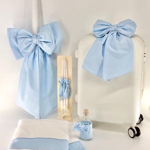 May include: A collection of baptism or christening gifts, featuring a white suitcase adorned with a light blue bow, a matching towel set, a candle with a large bow, and a small bottle with a bow. The items are in white and light blue.