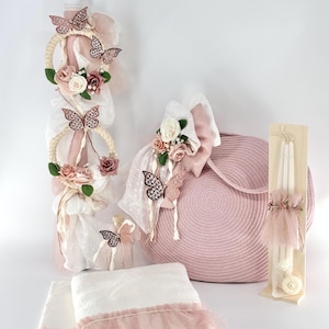 May include: A pink and white baby christening set featuring a candle, a basket, towels, and a small bottle. The candle is adorned with pink and white flowers, butterflies, and ribbons. The basket is made of woven straw and has a pink lining. The towels are white with pink trim. The bottle is clear glass with a pink butterfly decoration.