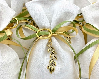 Puede incluir: Bolsas de regalo de lino blanco atadas con cintas verdes y doradas, aseguradas con anillos dorados y dijes de hojas. Las bolsas están llenas y recogidas en la parte superior, creando una apariencia suave y texturizada.