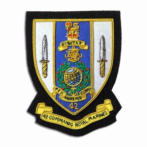 May include: A black, shield-shaped patch with gold trim and detailed embroidery. The design includes a blue central panel with a crest, two silver daggers, and the text "42 Commando Royal Marines" on a gold banner.