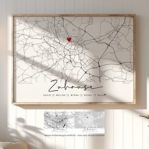 Op de afbeelding: Een zwart-witte kaartprint met een rood hart dat een locatie markeert. De kaart is ingelijst en heeft de tekst "Zuhause" en namen eronder. De tekst "Weitere Kartendesigns erhältlich - more map designs available" staat onderaan de afbeelding.