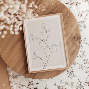 May include: A framed botanical line art print featuring a delicate floral stem with leaves and a trumpet-shaped flower. The artwork is in a light wood frame, resting on a round wooden table. The print's background is a soft beige, and the line art is black.