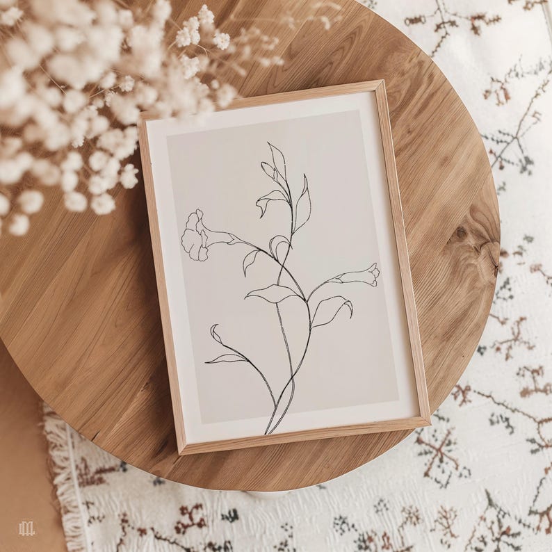 May include: A framed botanical line art print featuring a delicate floral stem with leaves and a trumpet-shaped flower. The artwork is in a light wood frame, resting on a round wooden table. The print's background is a soft beige, and the line art is black.