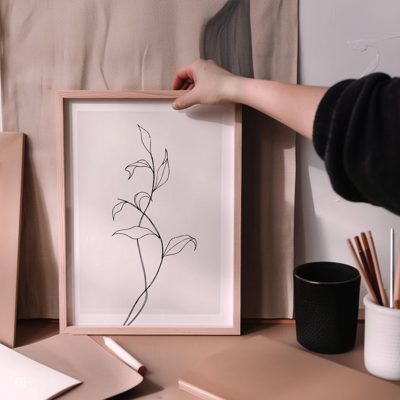 May include: A framed botanical art print featuring a minimalist line drawing of a plant with leaves. The artwork is in a light wood frame. The background includes a black textured cup, pencils, and a notebook on a neutral-toned surface.