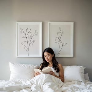 May include: Two framed botanical line art prints hang above a bed. A person is reading a book while sitting in bed, surrounded by white pillows and a white comforter. The art features black line drawings of flowers and leaves.