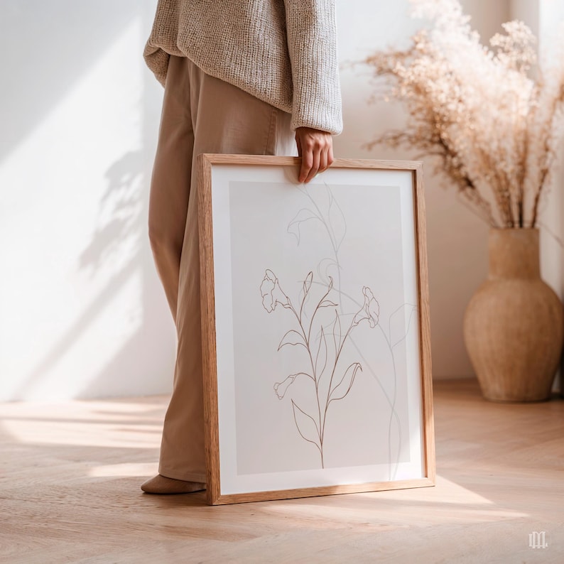 May include: A framed botanical art print featuring a minimalist line drawing of a flower with stems and leaves. The artwork is in a light brown wooden frame. The print is held by a person wearing a beige sweater and pants.