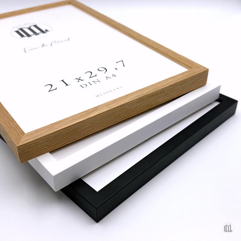 May include: Three picture frames stacked on top of each other. The top frame is light brown, the middle frame is white, and the bottom frame is black. The top frame has the text "21 x 29,7 DIN A4" and "Frame the Moment".