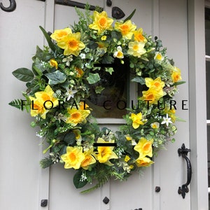 May include: A spring wreath with a circular design, showcasing bright yellow daffodils, green leaves, and small white blossoms. The wreath is mounted on a light grey door with black ironmongery and the text "FLORAL COUTURE".