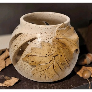 Puede incluir: Un portavelas de cerámica moteada de color blanquecino con un gran diseño de hoja en relieve en tonos marrones. El soporte redondo tiene varios recortes curvos y está decorado con pequeñas motas marrones.