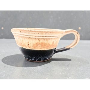 May include: A handmade ceramic cup with a handle. The cup features a two-tone design, with a beige upper half and a dark blue lower half. The handle is dark blue. The cup is likely used for drinking beverages.