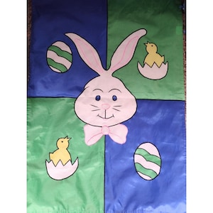May include: A green, blue, and pink Easter flag with a bunny rabbit, two chicks, and Easter eggs. The bunny has pink ears and a pink bow tie. The chicks are yellow and are in nests. The eggs are green and white.