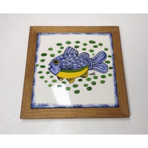 May include: A blue and yellow fish with green spots on a white ceramic tile framed in wood.