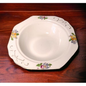 Puede incluir: Un tazón de cerámica blanca con un borde festoneado y un diseño floral. El tazón tiene un borde dorado y presenta flores amarillas, moradas y rosas.