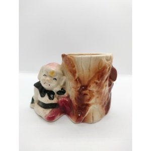 May include: A ceramic planter shaped like a tree stump with a small gnome sitting at the base. The gnome is painted in white, black, and red, and has a yellow star on its forehead. The tree stump is painted in brown and beige.