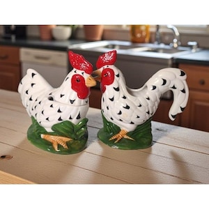 May include: Two ceramic chicken figurines. The chickens are white with black spots, red combs and wattles, and yellow beaks. They are perched on green bases with yellow feet. One has a lid.