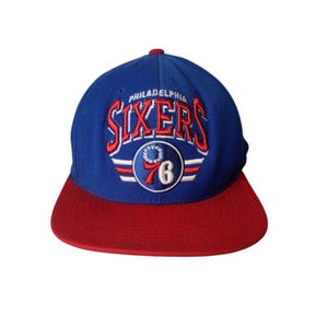 May include: A blue and red Philadelphia Sixers baseball cap. The cap features the team name "SIXERS" in red and white embroidery, along with a "76" emblem. The brim is red, and the crown is blue.