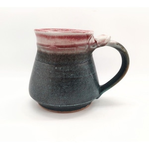 Może przedstawiać: Ręcznie robiony kubek ceramiczny z ciemnozieloną i fioletową glazurą. Kubek ma unikalny uchwyt i różowy rant.