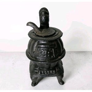 Op de afbeelding: Een zwarte gietijzeren miniatuur potkachel met een ronde behuizing, een klein deurtje aan de voorzijde met reliëfdetails, een schoorsteenpijp aan de bovenzijde en een gebogen handvat. De kachel staat op vier korte pootjes en heeft een textuur.