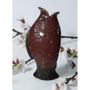May include: A maroon ceramic fish-shaped vase with a textured scale pattern. The vase has a dark brown base and accents. Delicate white and pink cherry blossom branches frame the vase, creating a decorative display.