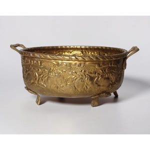 May include: A gold-colored, antique-style bowl with ornate floral and foliate relief patterns. The bowl has two handles and rests on three small feet. The rim features a rope-like design. A decorative piece for home decor.