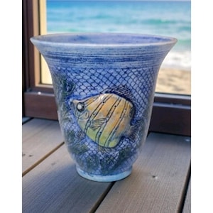 May include: A blue ceramic vase with a fish design. The vase features a yellow and brown fish, a blue net pattern, and green aquatic plants. The vase is on a wooden surface with a view of the ocean in the background.