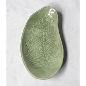May include: A light green ceramic leaf-shaped dish. The dish features a detailed embossed fern design in a slightly lighter shade of green. The overall shape is organic and the dish has a smooth, glossy finish. Ideal for serving or as a decorative piece.