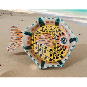 May include: A ceramic fish-shaped dish with a yellow interior and a white exterior. The dish features orange, teal, and black painted details. The fish tail is orange and white striped. The dish is on a sandy surface.
