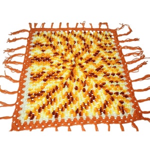 Puede incluir: Una manta cuadrada de crochet con un patrón geométrico en tonos naranja, amarillo y blanco. La manta tiene un borde de flecos.