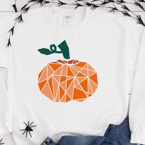 May include: White sweatshirt featuring a geometric orange pumpkin design. The pumpkin has a green stem and is made up of triangles.
