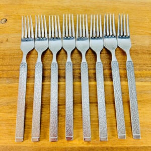 May include: A set of eight silver forks with textured handles. The forks are arranged in a row on a wooden surface. The tines of the forks are thin and evenly spaced.