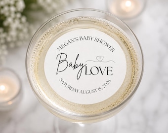 May include: A round white sticker with black text reading Megan's Baby Shower, Baby LOVE, and Saturday August 15, 2026, featuring a small heart graphic, placed on the surface of a clear glass filled with a pale yellow liquid beverage.