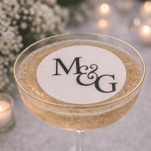 May include: A gold-rimmed coupe glass filled with a sparkling golden beverage. Floating on the surface is a circular white paper topper featuring the black printed letters M and G connected by an ampersand in a serif font.