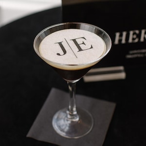 May include: A clear glass martini cocktail filled with a dark brown liquid, topped with a circular white paper garnish featuring the black printed letters J and E separated by a vertical line, resting on a square black napkin.