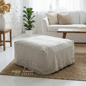 May include: A square, beige linen ottoman on a woven jute rug in a bright, white room. A white sofa with beige cushions and a wooden coffee table are in the background. A potted plant and a small wooden table are also visible.