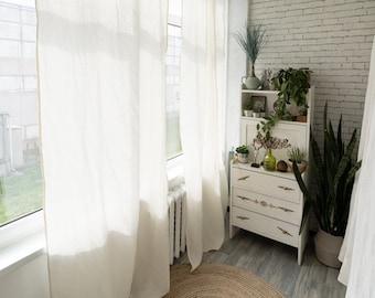 Puede incluir: Una habitación blanca con una pared de ladrillo blanco, una cómoda blanca con tres cajones y una alfombra redonda de yute. Hay plantas en la cómoda y en una maceta en el suelo. La habitación tiene cortinas blancas transparentes y una ventana con vista a la hierba verde.