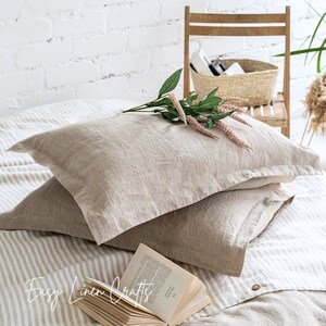 May include: Two beige linen pillowcases on a bed with a striped linen sheet and a book on top. The bed is in front of a white brick wall.