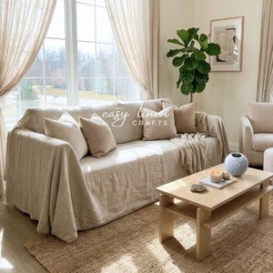 Op de afbeelding: Een woonkamer met een beige linnen bank, bijpassende kussens en een deken. De bank is bedekt met een linnen hoes. Een jute vloerkleed, een houten salontafel en transparante gordijnen zijn ook aanwezig. De tekst "easy linen crafts" is zichtbaar.