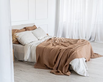 Può includere: Un copripiumino in lino marrone è drappeggiato su un letto in lino bianco con cuscini bianchi. Il letto si trova in una stanza bianca con pareti bianche e tende bianche trasparenti.