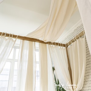 May include: A white linen canopy bed with a wooden frame and white curtains. The bed is decorated with a green plant and a white pillow.